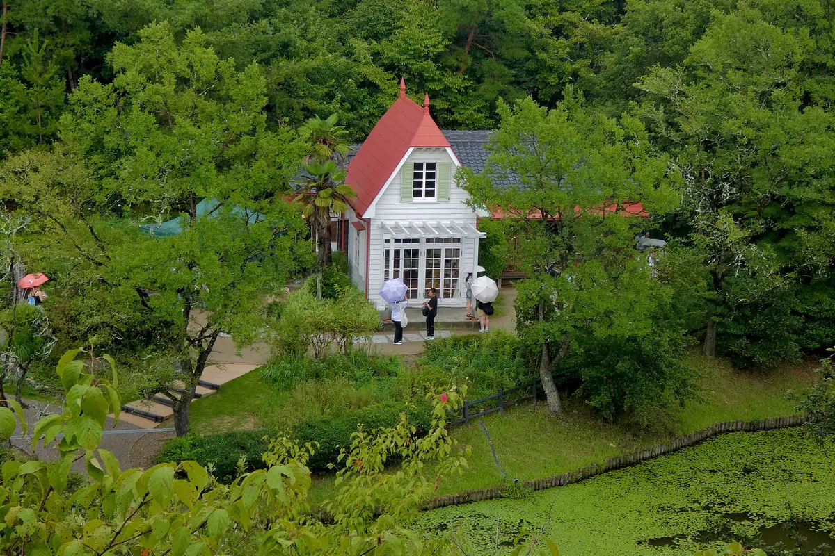 Satsuki and Mei's House, a white wooden house with a red pointed roof, seen from above among green trees, with visitors holding umbrellas at the door